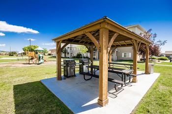 A wooden pavilion with a picnic table and chairs is situated in a grassy area.